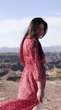 Rüzgar, kırmızı elbiseli, gülümseyen bir kadının saçlarında eser. Çöldeki bir kanyonun kenarında durur.