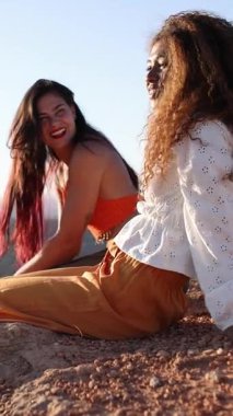 Kadın yoldaşlar, altın gün batımında kayalık uçurumun kenarında otururken keyifli bir anı paylaşıyorlar. Manzaranın ve karşılıklı bağlantının tadını çıkarıyorlar.