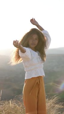 Altın gün batımı ışığında çıplak ayakla dans eden zarif kadın manken rüzgârlı saçları ve tasasız hareketleriyle dağlık arazide süzülüyor.