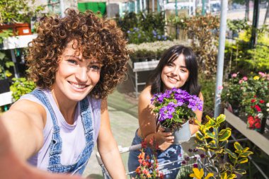 Bir bahçe merkezinde çalışan iki genç kadın çiçeklerle dolu bir alışveriş arabasıyla selfie çekiyorlar.