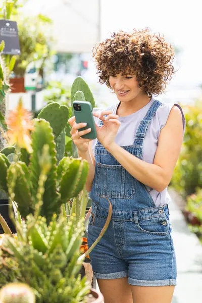 Gülümseyen kadın bahçıvan, serada ya da bahçe merkezinde bitki bilgilerini kontrol etmek için cep telefonu uygulaması kullanıyor