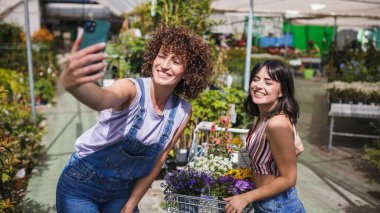 İki mutlu genç kadın, çiçeklerle dolu bir alışveriş arabasıyla bir bahçe merkezinde akıllı bir telefonla selfie çekiyor.