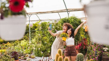 Bitki ve çiçeklerle dolu bir serada akıllı bir telefonla selfie çeken iki mutlu genç kadın işlerinin tadını çıkarıyorlar.