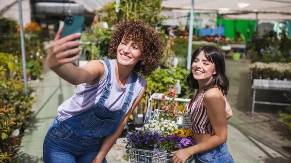 İki mutlu genç kadın, çiçeklerle dolu bir alışveriş arabasıyla bir bahçe merkezinde akıllı bir telefonla selfie çekiyor.