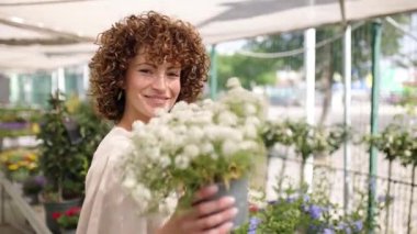 Profesyonel kadın bahçıvan çiçek açan bitkilerin yanında duruyor, elinde beyaz bir çiçek saksısı ve parlak seranın çalışma alanında içten bir gülümseme var.