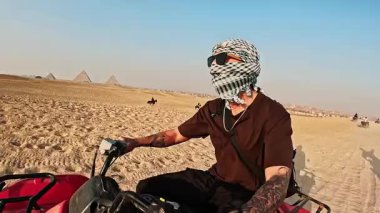 Güneş gözlüğü takan erkek turist ve ikonik giza piramitlerinin yanında dörtlü bisiklete binerek güneşli Mısır gökyüzü altında çöl macerası yaşıyor.