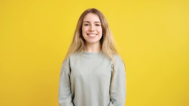 Uzun sarı saçlı, açık yeşil kazak giyen neşeli genç bir kadın canlı sarı bir arka planda poz veriyor ve gülümsüyor.