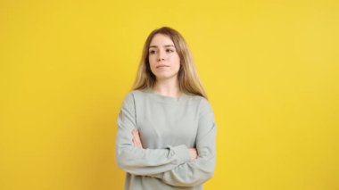 Çapraz kollu ve ciddi bir yüz ifadesi olan genç bir kadın, parlak sarı bir arka planda izole edilmiş.
