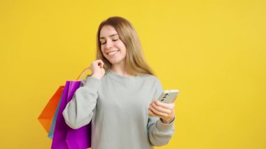 Akıllı telefonları kontrol ederken gülümseyen genç bir bayan. Elinde parlak sarı stüdyo geçmişine karşı çoklu alışveriş torbaları var. Modern dijital alışveriş deneyimleri sergiliyor.