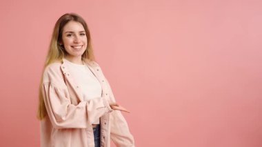 Genç bir kadın sevinçle el kol hareketi yapıyor ve açık pembe bir zemin üzerinde boş bir alana işaret ediyor, sunumlar veya ürünler sergiliyor.