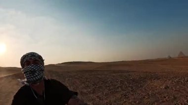 Genç erkek, kumlu çöl arazisinde ikonik giza piramitleriyle bisiklete biniyor altın gün batımına karşı siluetlenmiş, Mısır 'da maceralı bir yolculuk anı sergiliyor.