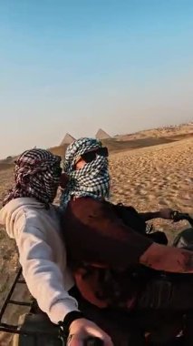 İkonik Giza Piramitleri yakınlarındaki çöl manzarasında turistler dörtlü bisikletlerle kumlu arazide geziniyor, tarihi Mısırlılar arasında maceralı bir kültürel yolculuk yaşıyor.