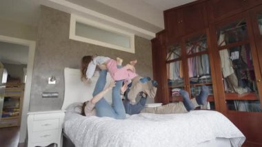Sevgi dolu ebeveynler, yatak odasında dinlenirken ayaklarını kullanarak neşeli bir şekilde kızlarını kaldırır, gerçek bir aile sevinci ve bağ anını paylaşırlar.