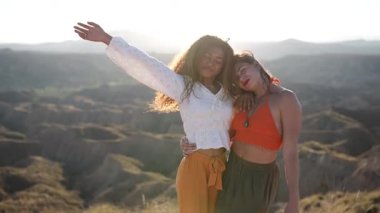 Kadın arkadaşlar yakınlaşma anını paylaşıyorlar, çöl manzarasıyla birlikte duruyorlar, altın gün batımında neşeli bir şekilde birbirlerine sarılıyorlar.