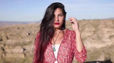 Dalgalı kırmızı elbisesiyle kendinden emin bir kadın kayalık çöl kanyonunun ortasında dikiliyor, dramatik manzaraya karşı zarif bir stil sergiliyor mavi gökyüzü ve dağlık arazisiyle