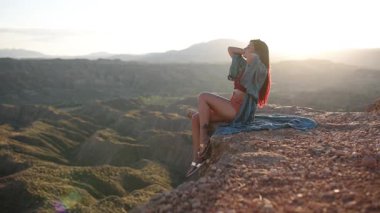 Kayalık uçurumun kenarında duran, altın gün batımında şık bir bornoz giyen, dramatik çöl manzarasını aydınlatan özgüvenli bir manken.