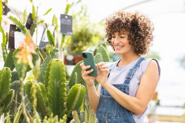 Çeşitli kaktüsler ve yemyeşil bitkilerle dolu canlı bir serada akıllı telefon kullanarak gülümseyen mutlu kadın bahçıvan.