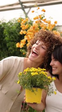 İki mutlu bayan çiçekçi akıllı bir telefonla selfie çekiyorlar ve bir serada sarı bir saksı çiçeğiyle poz veriyorlar. Dikey