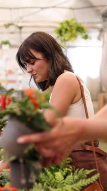 Renkli çiçeklere göz atan, bahçecilik sezonunda güneşli serada ya da bahçe merkezinde dikilirken özenle bitkileri seçen bir kadın. Dikey