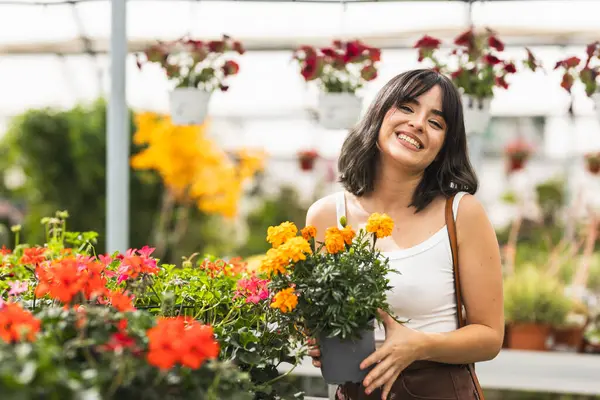 Canlı bir serada renkli bir çiçek dizisiyle çevrili iken canlı bir kadife çiçeği tutan mutlu genç kadın.