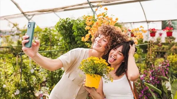 Gülümseyen genç bayan arkadaşlar birlikte selfie çekerken ellerinde sarı saksılı çiçeklerle renkli serada canlı çiçeklerle eğleniyorlar.