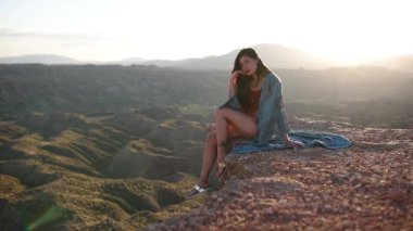 Günbatımında çöl kanyonu manzarasına bakan bir uçuruma poz veren mayo ve açık plaj bornozu giyen genç bir kadın.