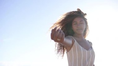 Genç bir kadın dans ederek özgürlüğünü ve neşesini ifade eder açık mavi gökyüzü ile güneşli bir ortamda, rüzgar hafifçe saçlarını savurur.