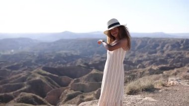 Uçan elbiseli ve şapkalı gülümseyen kadın uçurumun kenarında neşeyle dans ediyor. Engebeli kanyon manzarası, berrak mavi gökyüzünün altına kadar uzanıyor.