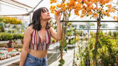 Genç bir kadın renkli bir serada yemyeşil çiçeklerle çevriliyken canlı turuncu begonvillerin enfes aromasının tadını çıkarıyor.