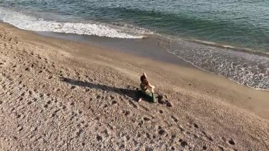 Sakin kadın yoga uygulayıcısı kumlu kıyı şeridinde meditasyonunu gerçekleştiriyor, dikkatli fiziksel hareketlerle nazikçe dalgaların etrafında hareket ediyor.