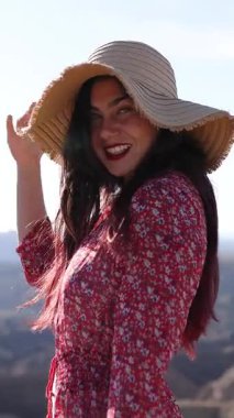 Canlı kırmızı çiçekli elbise ve hasır şapka giyen şık bir manken, sıcak güneş ışığıyla manzaralı doğal manzaraya karşı özgüvenle duruyor.