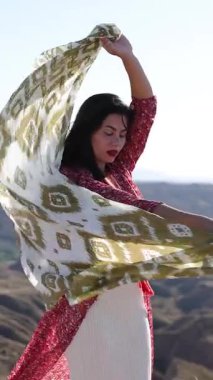 Zarif manken, dalgalı eşarpla zarif bir şekilde dans ediyor. Sıcak güneş ışığıyla yıkanmış dramatik dağ manzarasına karşı özgürce hareket ediyor.