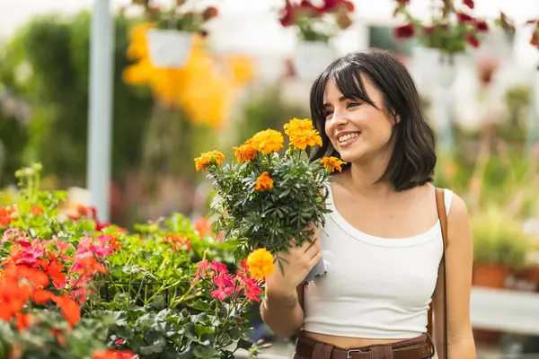Beyaz mahsul bluzunda gülümseyen genç bir kadın güneşli bir günde seranın içinde canlı sarı kadife çiçekleri tutuyor.