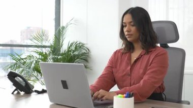 Modern ofis masasında dizüstü bilgisayarla çalışan profesyonel iş kadını, iş günü kameraya bakarken gülümsüyor.