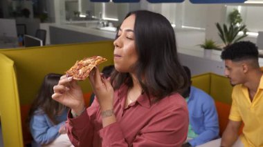 Genç profesyonel ofis molasında bir dilim pizzanın tadını çıkarıyor, rahat bir şekilde gülümsüyor ve günlük iş ortamında oturuyor.