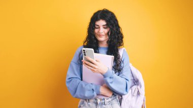 Sırt çantası ve not defteri olan genç bir kadın canlı sarı arka planda akıllı telefon kullanıyor, modern eğitim ve teknolojiyi temsil ediyor.