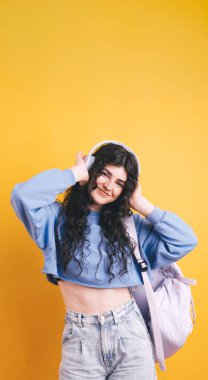 Canlı sarı arka planda müzik ve sırt çantasıyla müzik dinleyen gülümseyen öğrenci, eğitimi, gençliği ve teknolojiyi temsil ediyor.