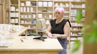 Professional female artist carefully sculpting detailed clay face sculpture, working with precision in well-organized pottery studio workspace
