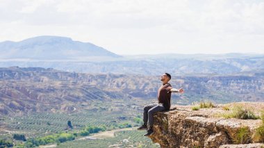 Bir uçurumun kenarında oturan genç adam kollarını açmış İspanya 'daki görkemli Gorafe Çölü manzarasını seyrediyor.