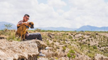 Genç adam boksör köpeğini okşarken bir uçurumun kenarında oturmuş Gorafe, İspanya 'nın çöl manzarasını seyrediyor.