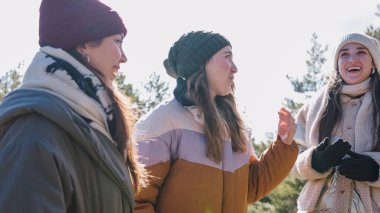 Üç neşeli kadın kış kıyafetleri giyip güneşli bir kış gününde sohbet ediyorlar ve el kol hareketi yapıyorlar.
