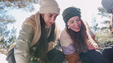 İki dişi yürüyüşçü, güneşli bir ormanda kış yürüyüşü sırasında karda bir şeyi inceliyor ve çömeliyor.