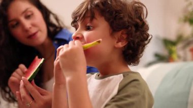 Genç bir çocuk, parlak oturma odasında güzel bir anneyle oturup sağlıklı atıştırmalık ve birliktelik dolu bir aile anını paylaşırken sulu karpuz diliminin tadını çıkarıyor.