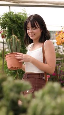 Sera işçisi terracotta kaktüs tenceresini dikkatle tutuyor, bahçenin içindeki bitkileri dikkatlice tarıyor ve profesyonel davranışlarına odaklanıyor. Dikey