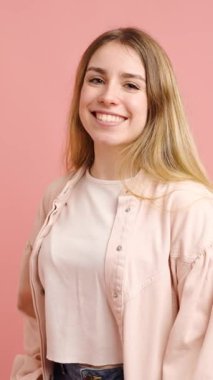 Genç bir kadın, dinamik yüz ifadeleriyle mutluluk ve neşe ifade ederken yumuşak pembe arka planda poz veriyor.