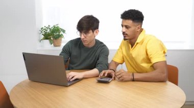 İki genç iş adamı, modern bir ofiste, yuvarlak bir masada dizüstü bilgisayar ve hesap makinesi kullanarak birlikte çalışıyorlar.