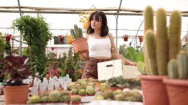 Bitkilerle dolu masalarda yürürken saksıda kaktüs ve tahta kutu taşıyan bir sera işçisi.