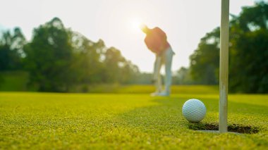 Golfçü topu yeşil golfün üstüne koyuyor, mercek akşam vakti parlıyor. Golfçü vuruşu uzun süre deliğe soktuktan sonra kazanır..                                 