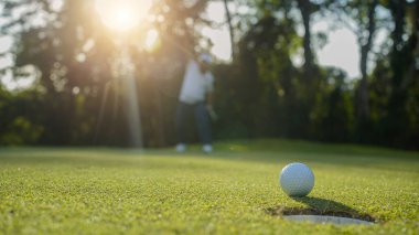 Golfçü topu yeşil golfün üstüne koyuyor, mercek akşam vakti parlıyor. Golfçü vuruşu uzun süre deliğe soktuktan sonra kazanır..                                