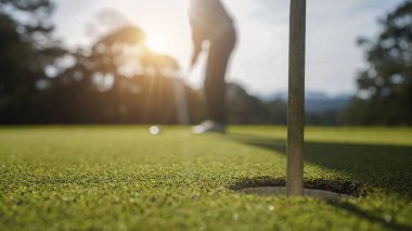 Golfçü topu yeşil golfün üstüne koyuyor, mercek akşam vakti parlıyor. Golfçü vuruşu uzun süre deliğe soktuktan sonra kazanır..                                